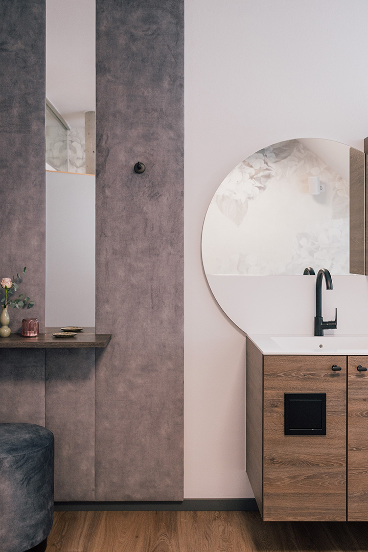 Moderner Waschtisch mit rundem Spiegel, schwarzem Wasserhahn und Holzschränken neben einer grauen gepolsterten Wand mit einem kleinen Tisch und einem Blumenarrangement.
