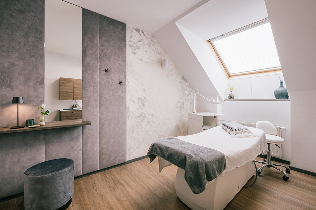 Heller Behandlungsraum mit Massagebett, Stuhl, großem Fenster und moderner Einrichtung mit Holzboden.
