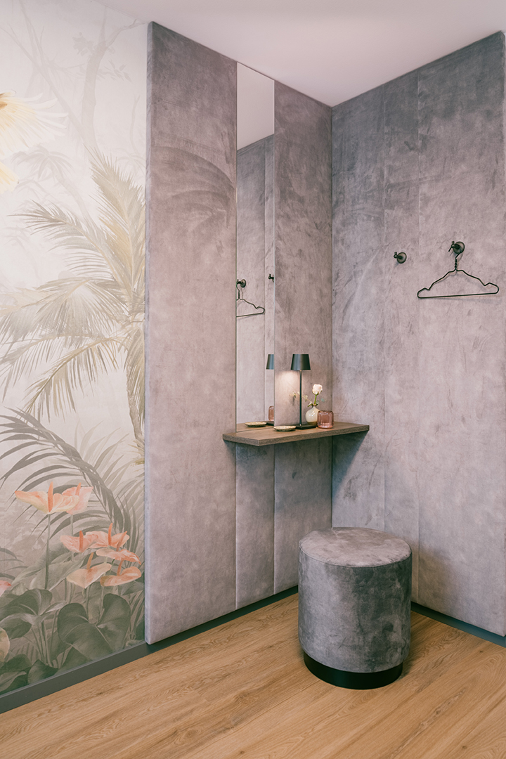 Eleganter Ankleidebereich mit grauem samtigen Hocker, eingebautem Schminktisch mit Tischlampe und Blume, umgeben von samtbezogenen grauen Wänden und einer Wand mit botanischem Muster.