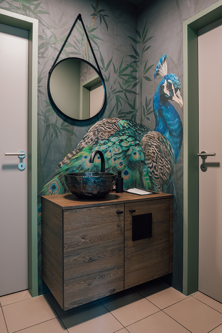 Kleines Badezimmer mit Holzmöbel, rundem Wandspiegel und Wandbild eines Pfaues mit blauen und grünen Federn.