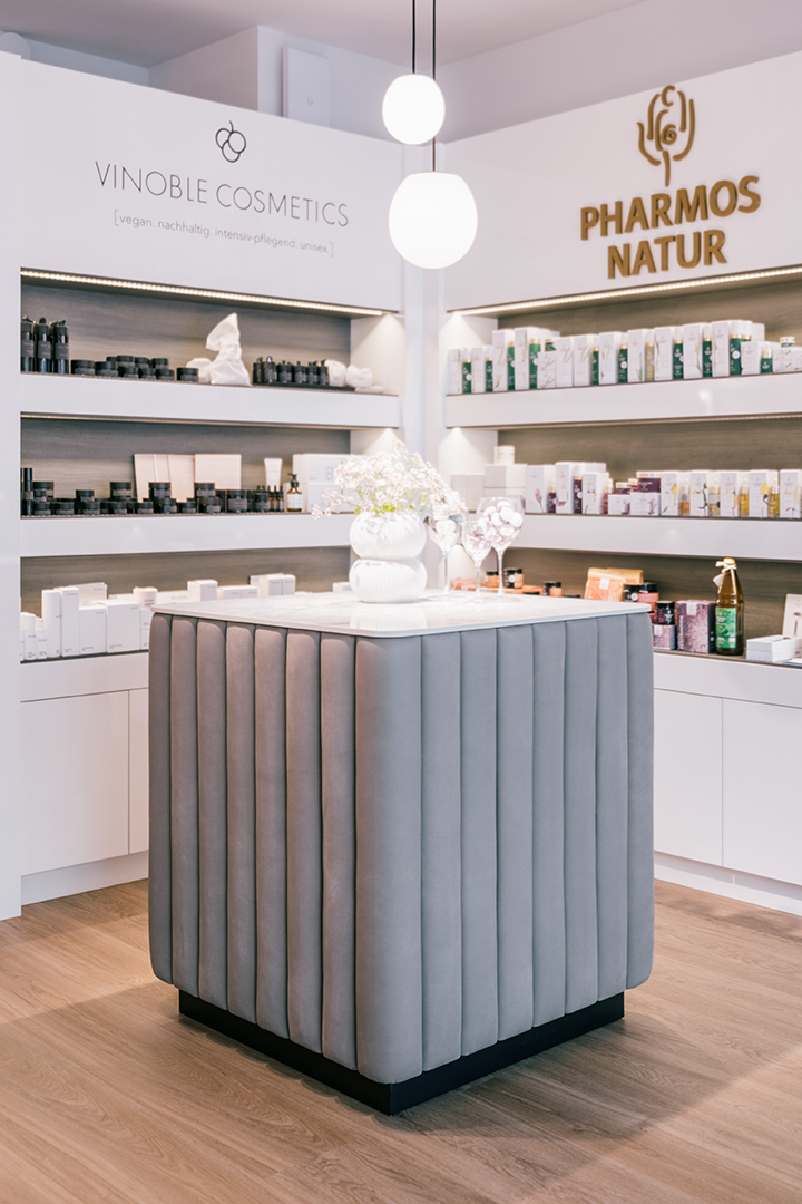 Moderne Kosmetikladen-Innenansicht mit einem stilvollen grauen gepolsterten Displaytisch und hell erleuchteten Regalen mit Vinoble Cosmetics und Pharmos Natur Produkten.