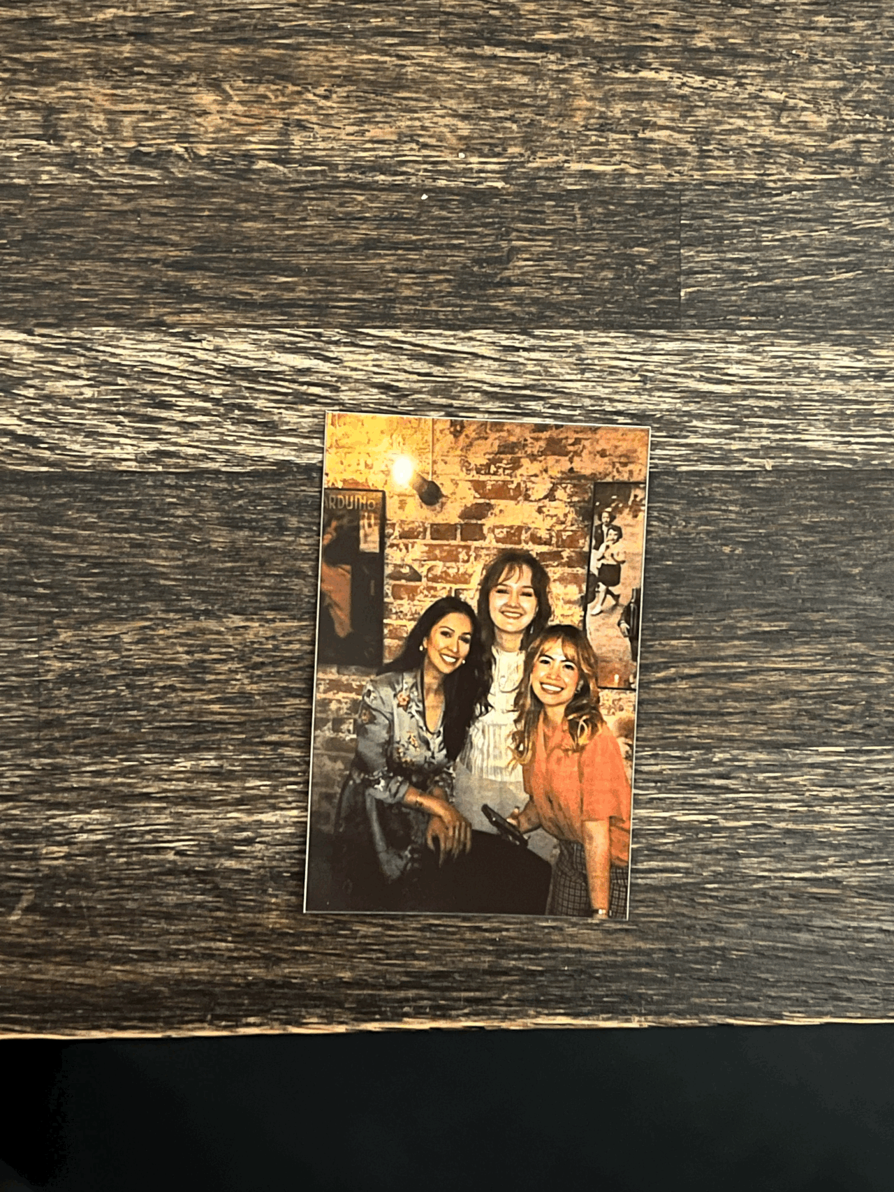 Photograph of three smiling women posing together against a brick wall backdrop on a wooden table.