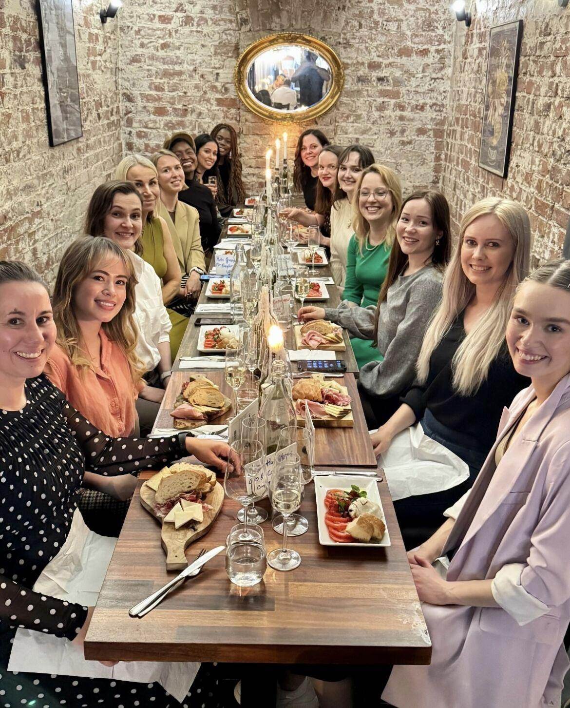 Group of smiling women seated along both sides of a long wooden table with plates of food and wine glasses in a room with exposed brick walls and a mirror.