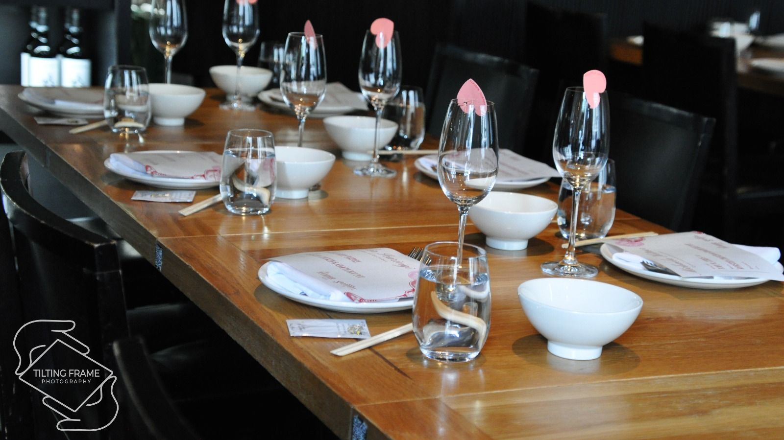 Wooden dining table set with white bowls, water glasses, wine glasses, chopsticks, and plates with folded napkins and menus in a restaurant.