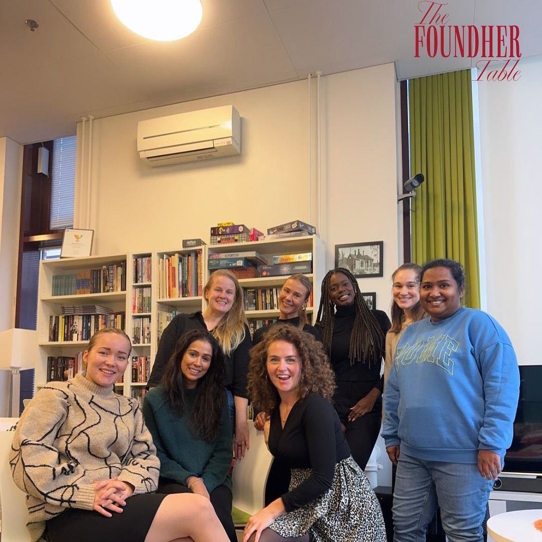 Group of eight diverse women smiling and posing together in a cozy room with bookshelves and a large window.
