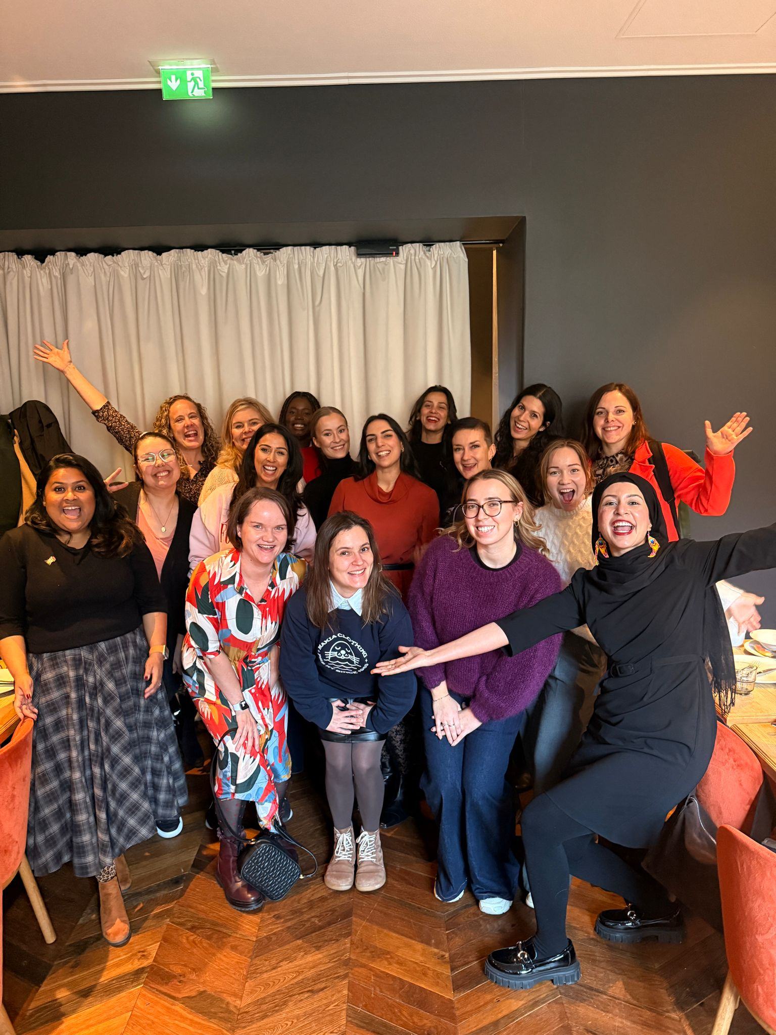 Group of diverse women smiling and posing enthusiastically indoors on a wooden floor with light-colored curtains in the background.