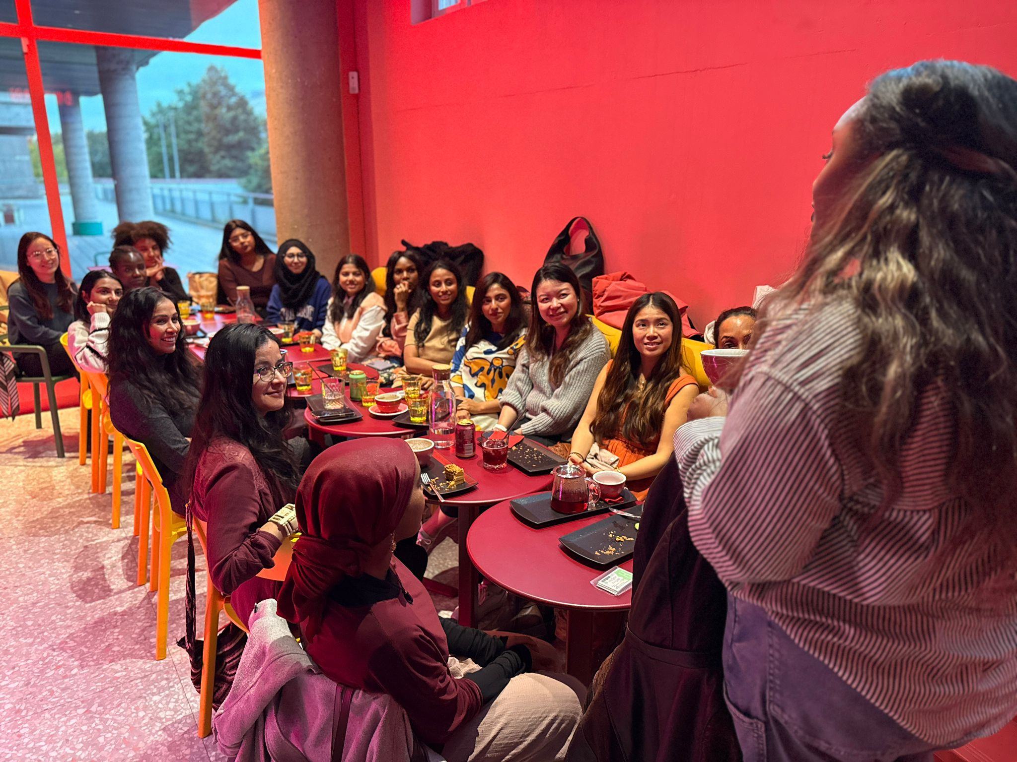Group of diverse women seated around red tables in a warmly lit room, attentively listening to a woman standing and speaking.