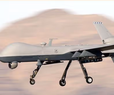 Gray military drone flying with landing gear extended against a blurred mountainous background.