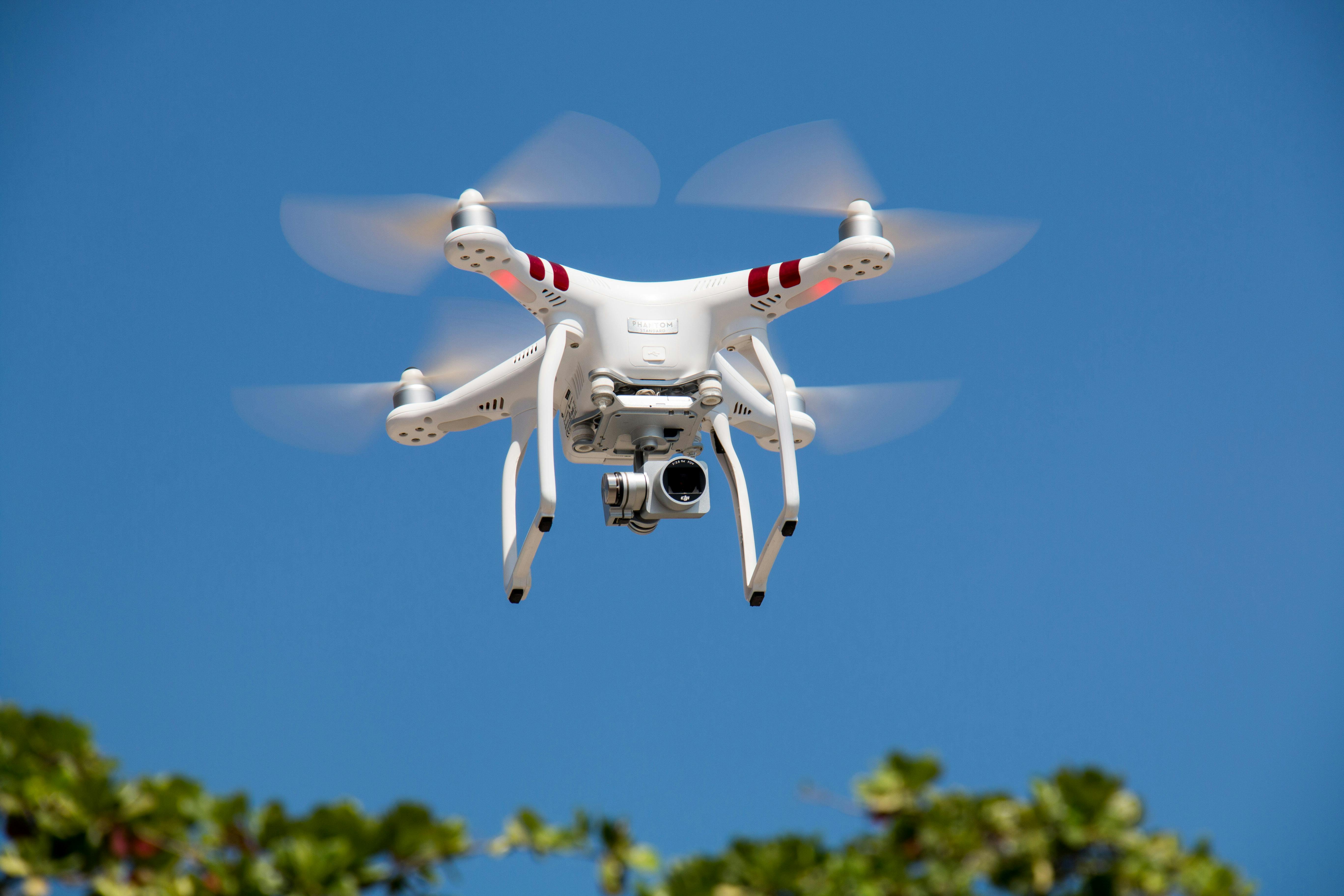 White quadcopter drone flying against a clear blue sky with blurred spinning propellers.