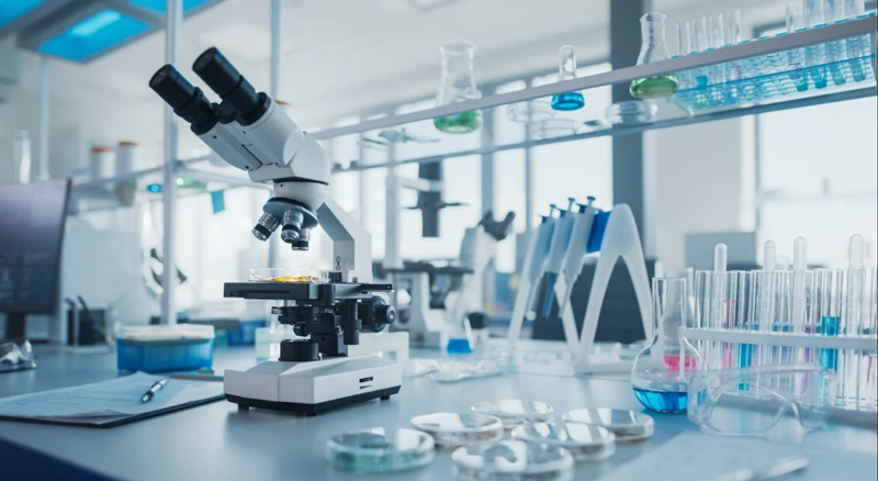 Close-up of a microscope on a laboratory table surrounded by petri dishes, test tubes with colorful liquids, and lab equipment in a bright modern lab.