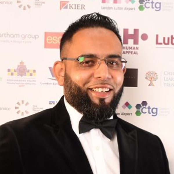 Formal event participant wears black tuxedo with glasses and various sponsor logos displayed.