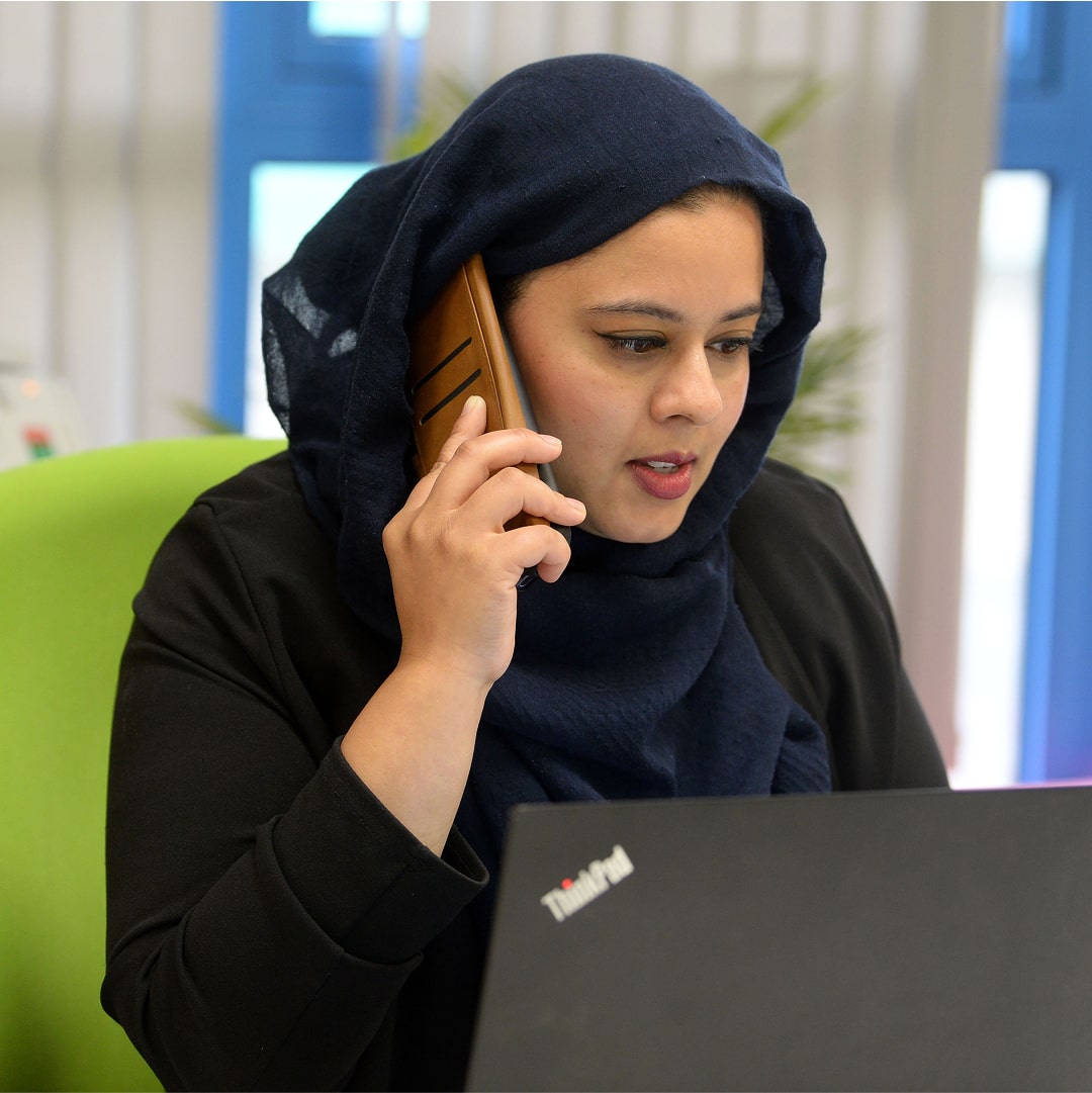 Woman in hijab using ThinkPad laptop, taking phone call in modern workspace setting.