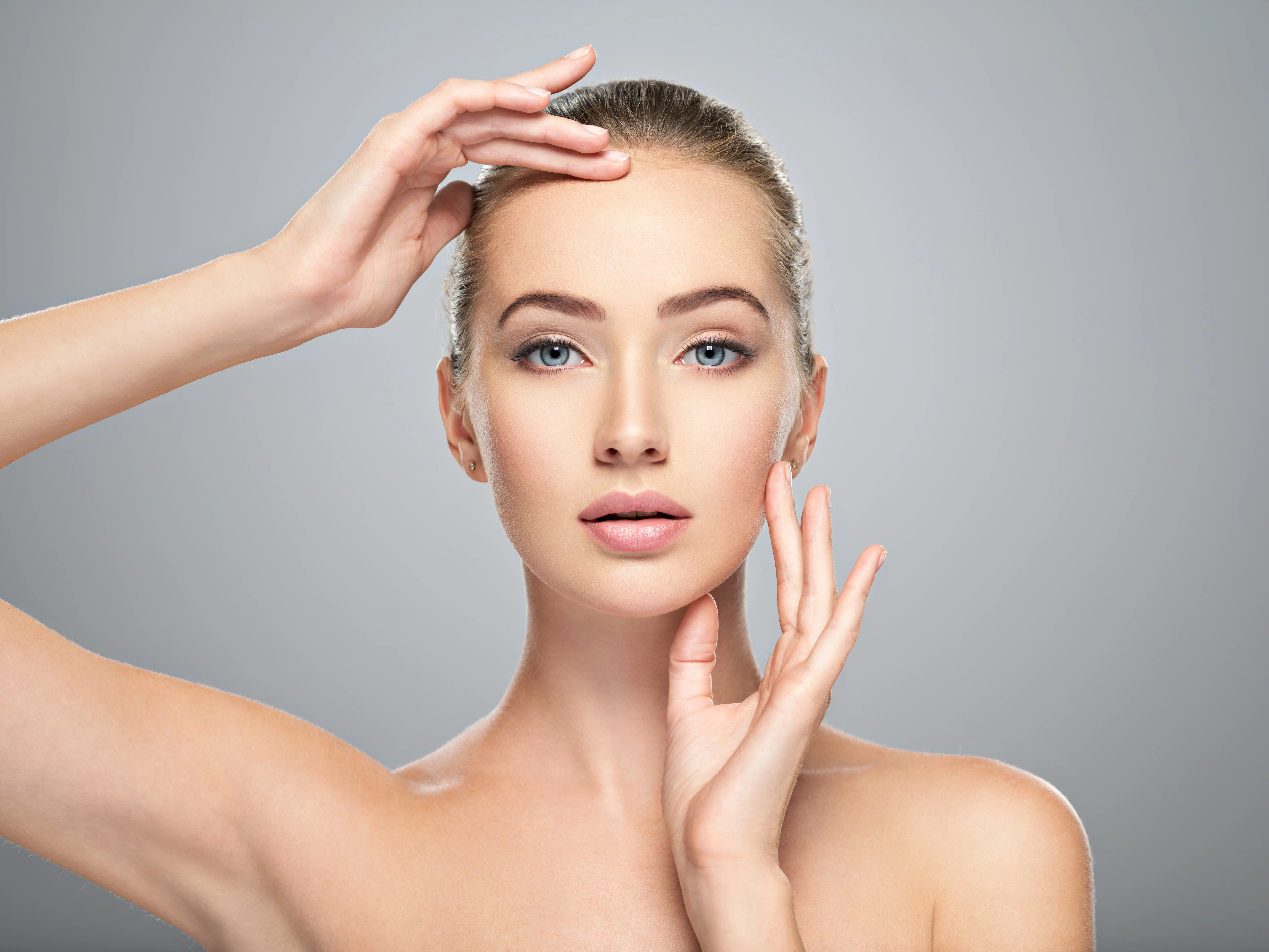 Close-up of a woman with clear skin gently touching her forehead and cheek against a gray background.