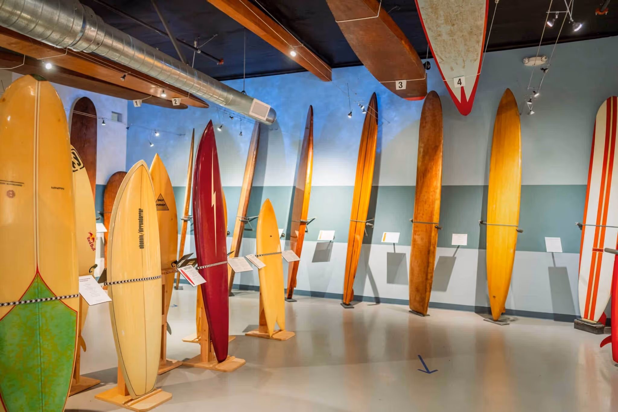 Exhibition room displaying various vintage wooden surfboards arranged on stands against a blue and teal wall.