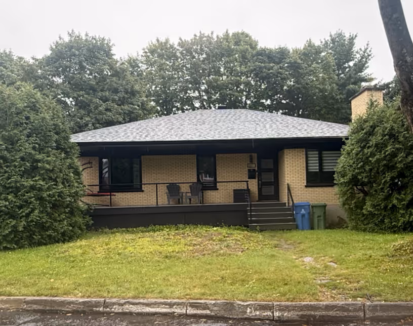 Maison en brique beige avec un porche, deux chaises, un escalier et des poubelles bleue et verte sur une pelouse verte bordée d'arbres.