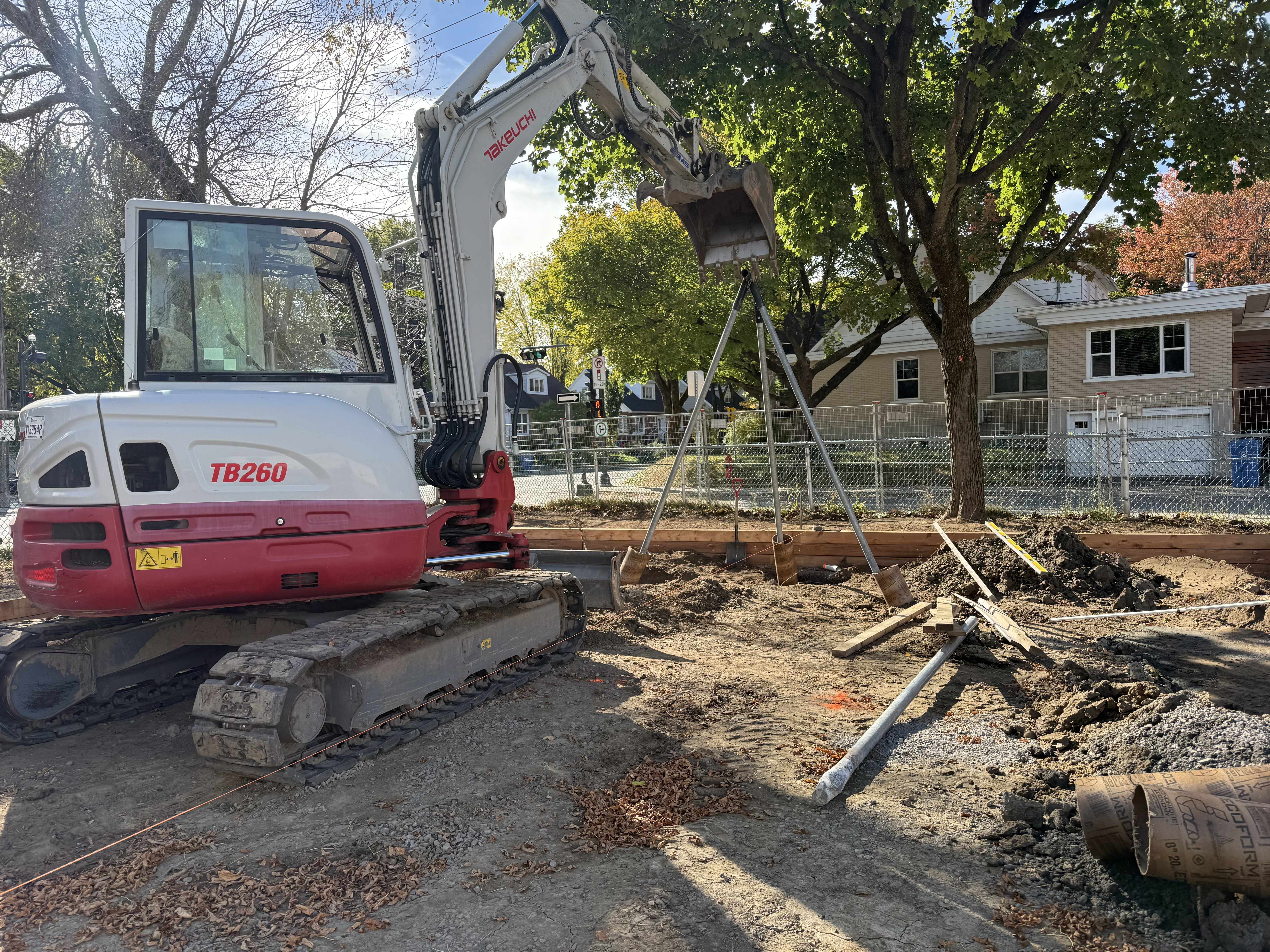 Petite colline herbeuse aménagée avec des rochers et deux jeunes arbres, bordée par un bâtiment en brique et une surface asphaltée humide.