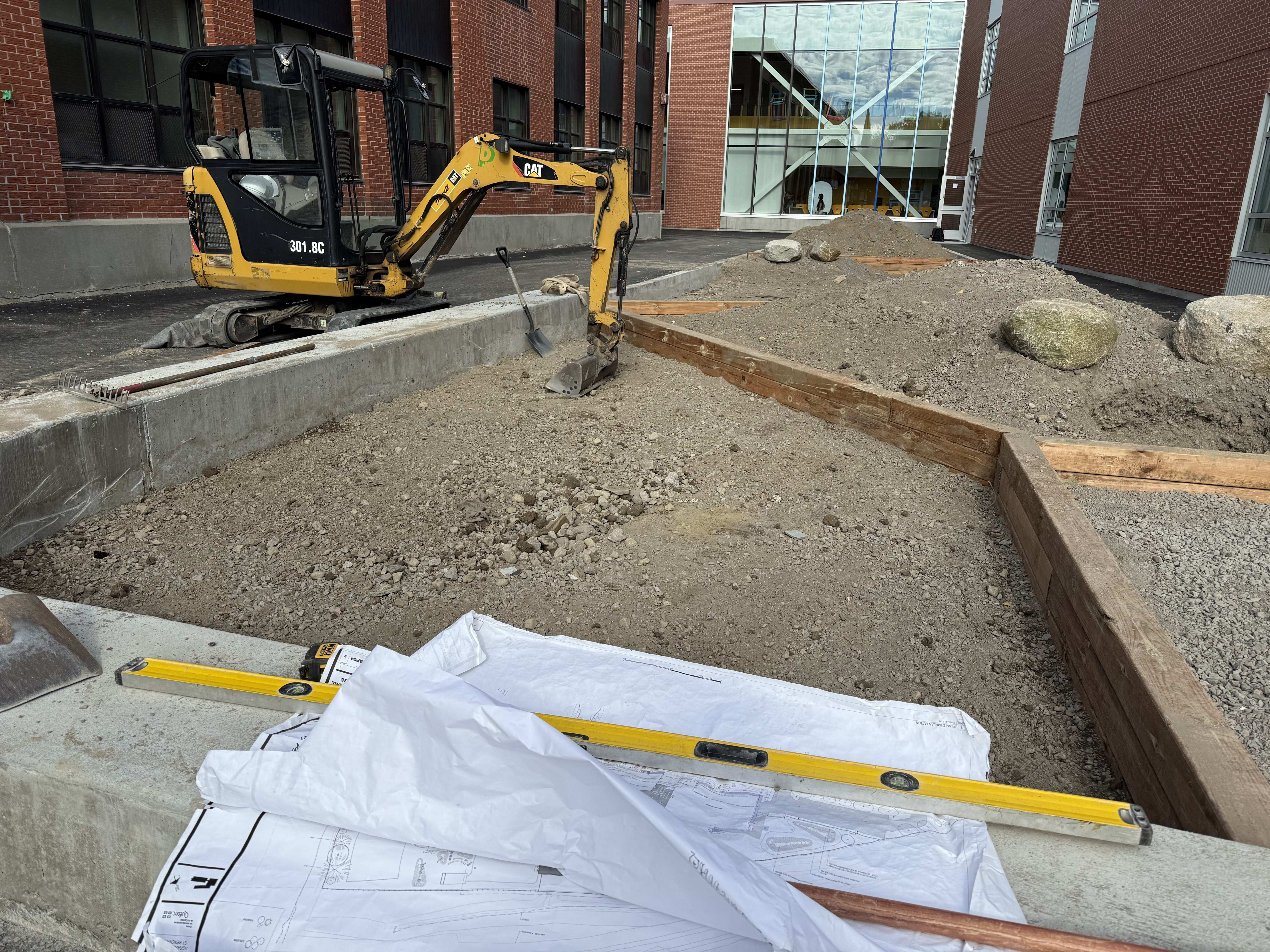 Mur de soutènement en blocs de béton gris le long d'un trottoir avec un terrain en pente et des arbustes au-dessus, sous un ciel nuageux.