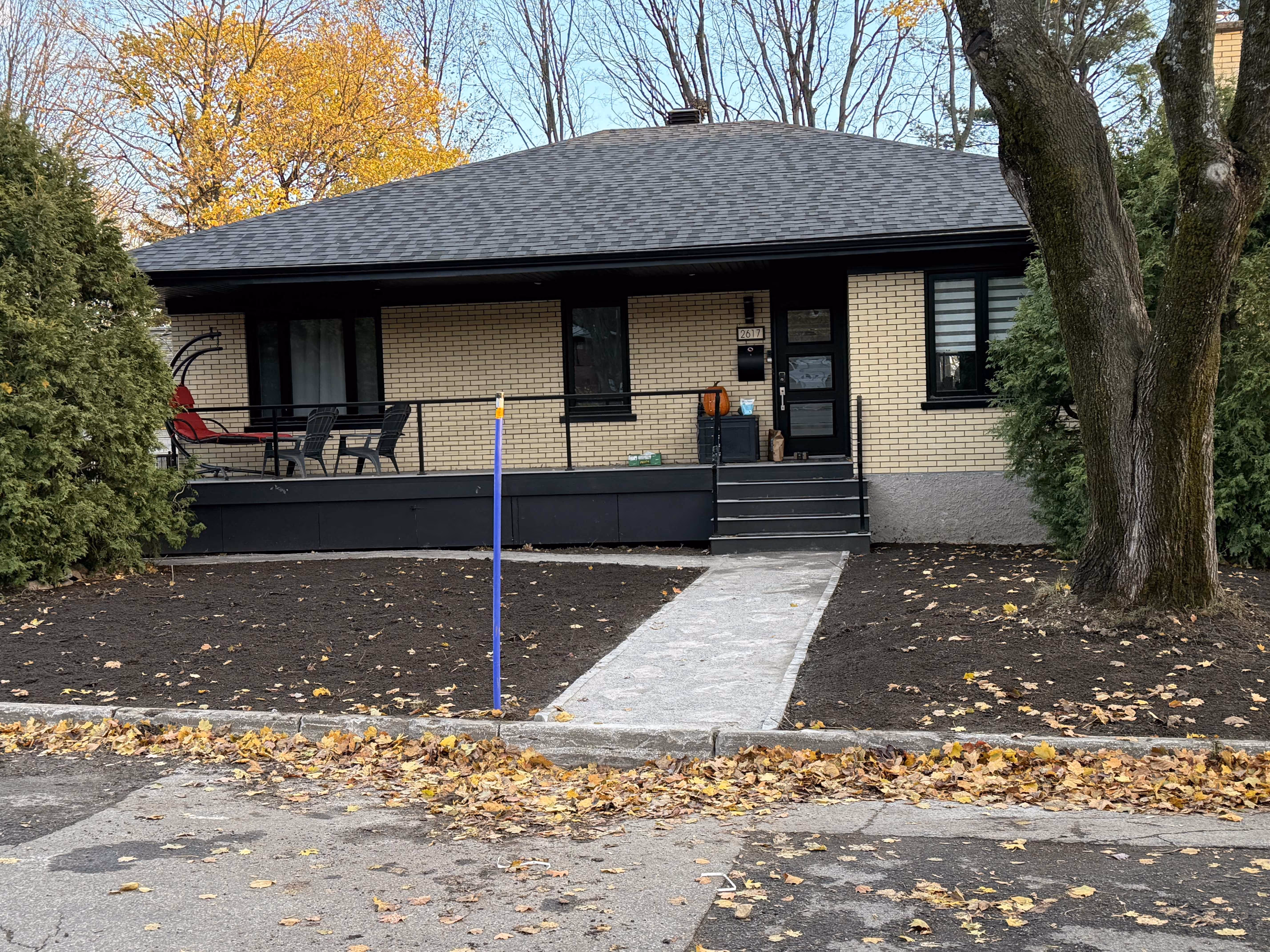 Maison avec façade en briques jaunes, terrasse noire avec chaises, allée pavée et sol fraîchement nivelé sans végétation.