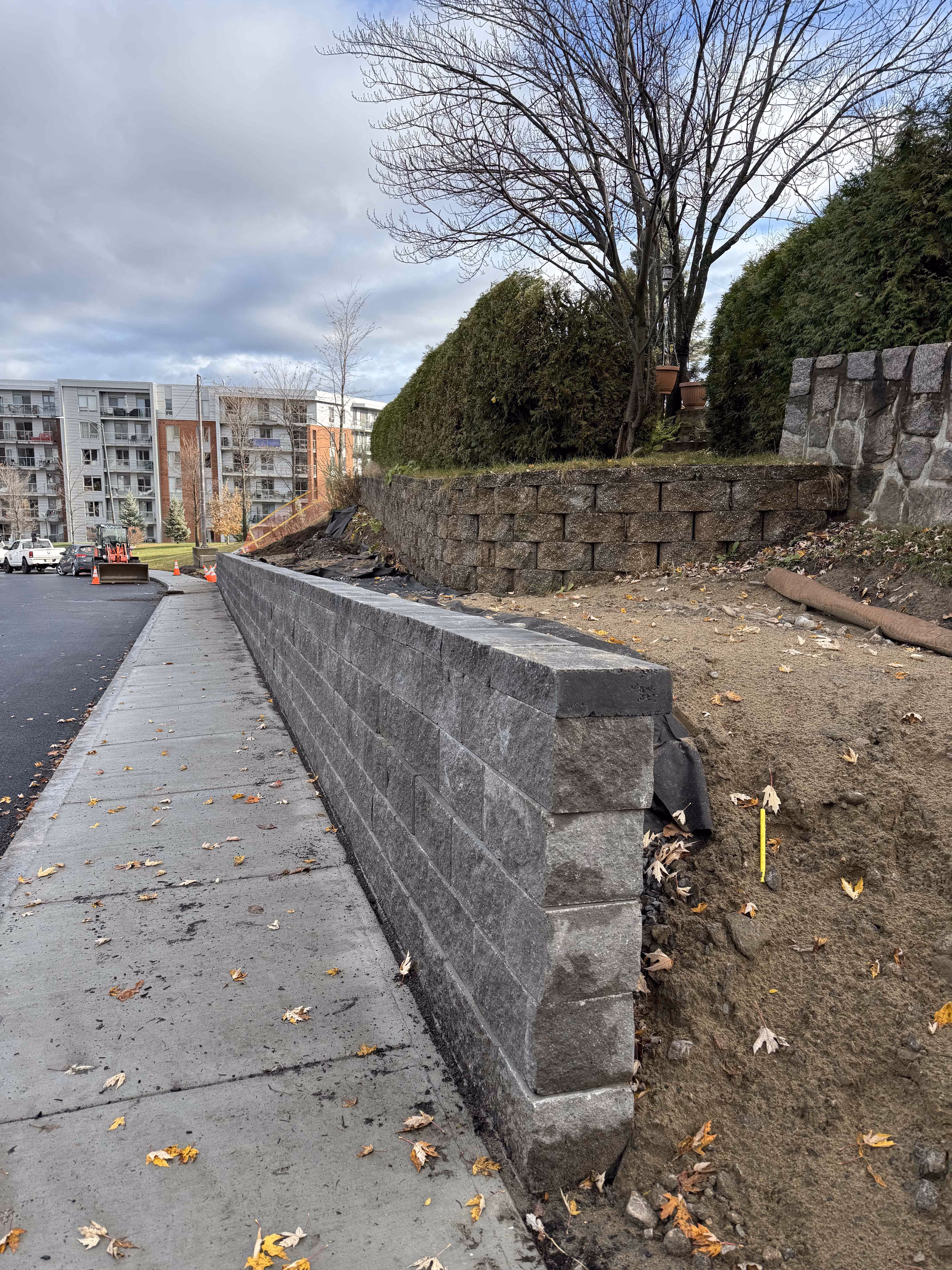 Mini-pelle sur un chantier avec plans et niveau à bulle posés sur un mur en béton devant un sol en gravier partiellement encadré par des madriers en bois.
