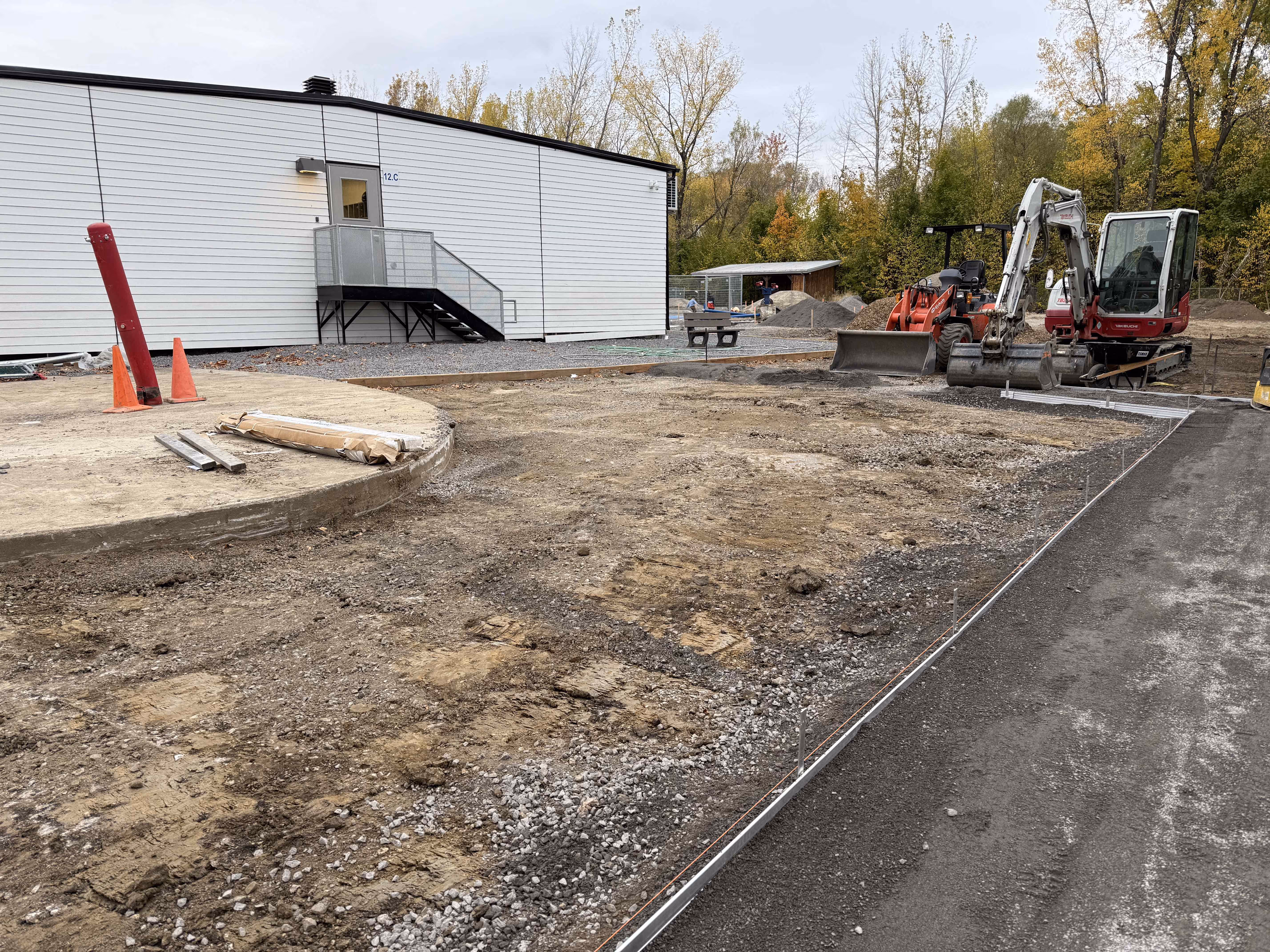 Zone de construction avec terrassement, engins de chantier et bâtiment blanc à l'arrière-plan en automne.