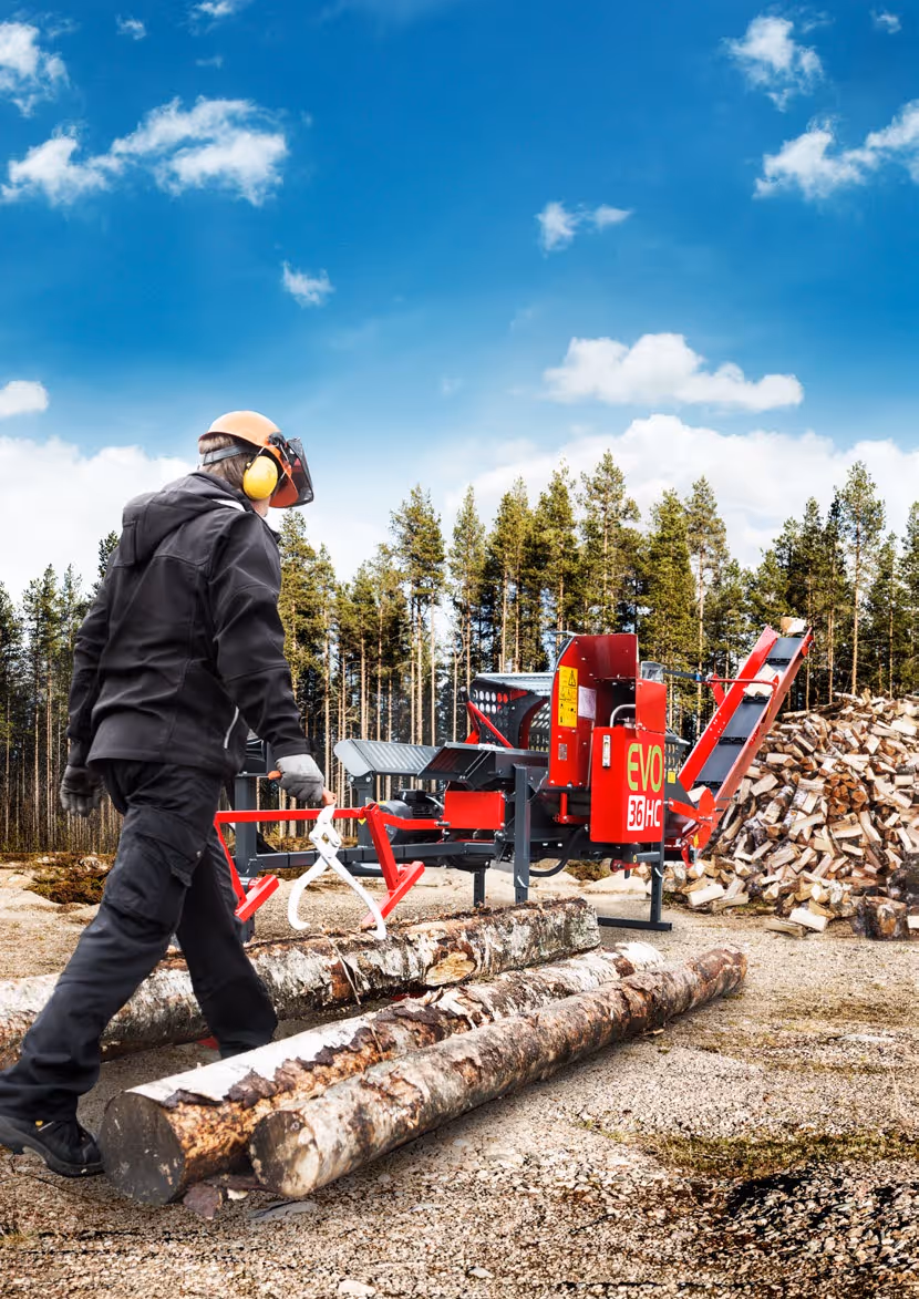 Opérateur en tenue de protection marchant vers une machine de découpe de bois rouge à l'extérieur près d'un tas de bûches et de troncs d'arbres.