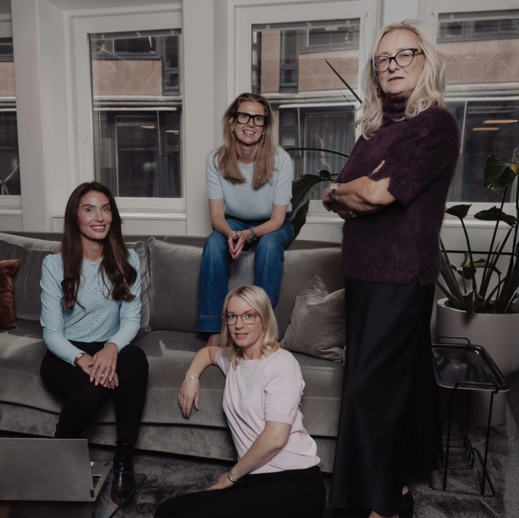 Fyra kvinnor i vardagsrumsmiljö, tre sitter på en grå soffa och en står bredvid, alla tittar mot kameran.