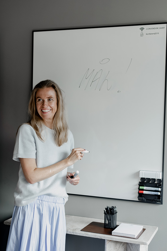 Kvinna med långt blont hår som ler och håller en whiteboard-pennor framför en whiteboard med ordet 'MÅH!' skrivet på den.