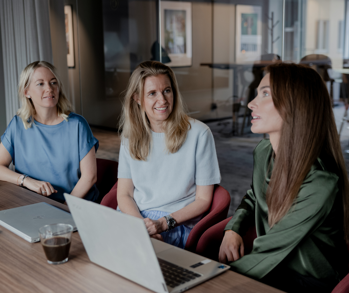 Tre kvinnor sitter runt ett bord med bärbara datorer och samtalar i ett modernt kontorsutrymme.