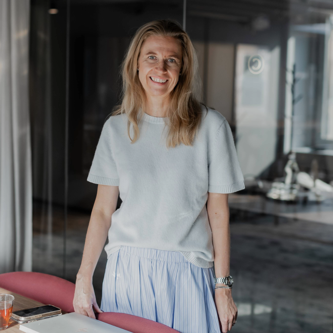 Fyra kvinnor i vardagsrumsmiljö, tre sitter på en grå soffa och en står bredvid, alla tittar mot kameran.