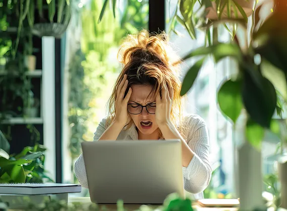 Frustrated woman with glasses holding her head while looking at a laptop in a bright room filled with green plants.