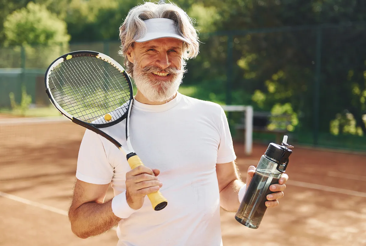 Smiling older man on tennis court holding a tennis racket and water bottle.