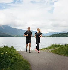 A man and woman jogging together on a paved path by a lake with mountains in the background under a partly cloudy sky.