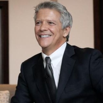 Smiling middle-aged man with gray hair in a black suit and tie sitting indoors.
