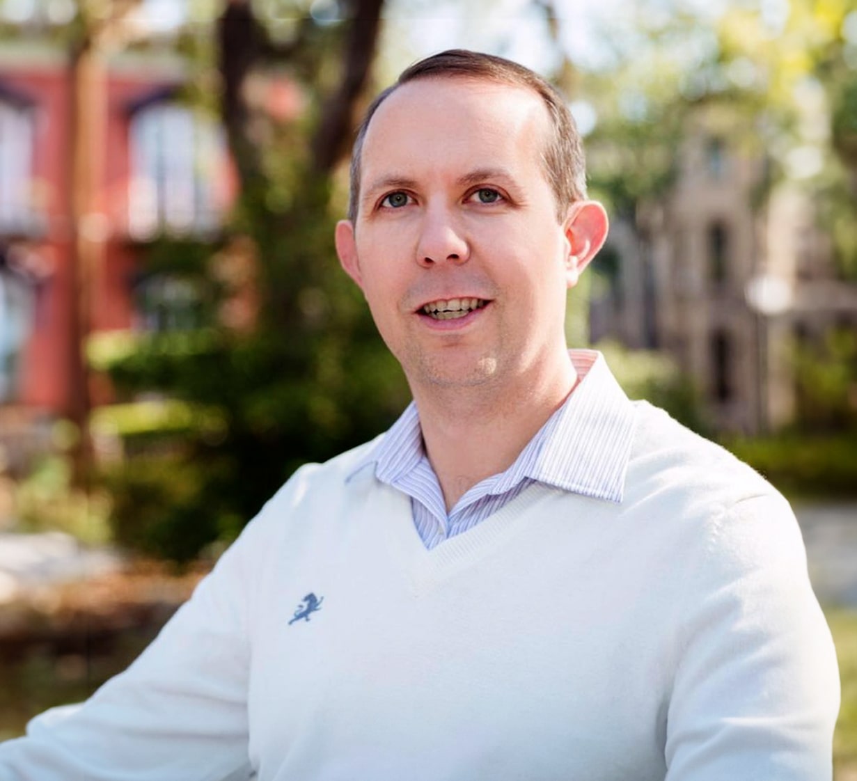 Smiling man wearing a white sweater and striped shirt outdoors with blurred trees and buildings in the background.