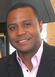 Smiling man with short hair wearing a black suit jacket over a light pink shirt.
