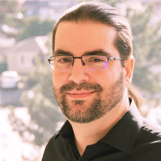 Man with dark hair tied back, beard, glasses, and black shirt smiling with a blurred outdoor background.
