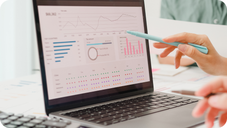 Person pointing at a laptop screen showing various colorful data charts and graphs including bar charts, a donut chart, and a line graph.