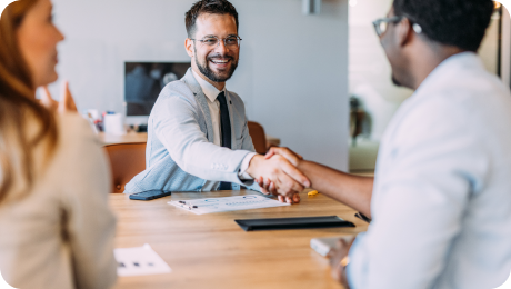 Man in glasses and light gray blazer shaking hands with another person across a table in an office setting, while a woman watches.