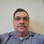Middle-aged man with glasses and short dark hair wearing a blue shirt, looking at the camera against a plain light background.