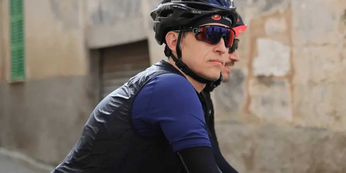 Two cyclists in helmets and sunglasses riding side by side near a stone wall.