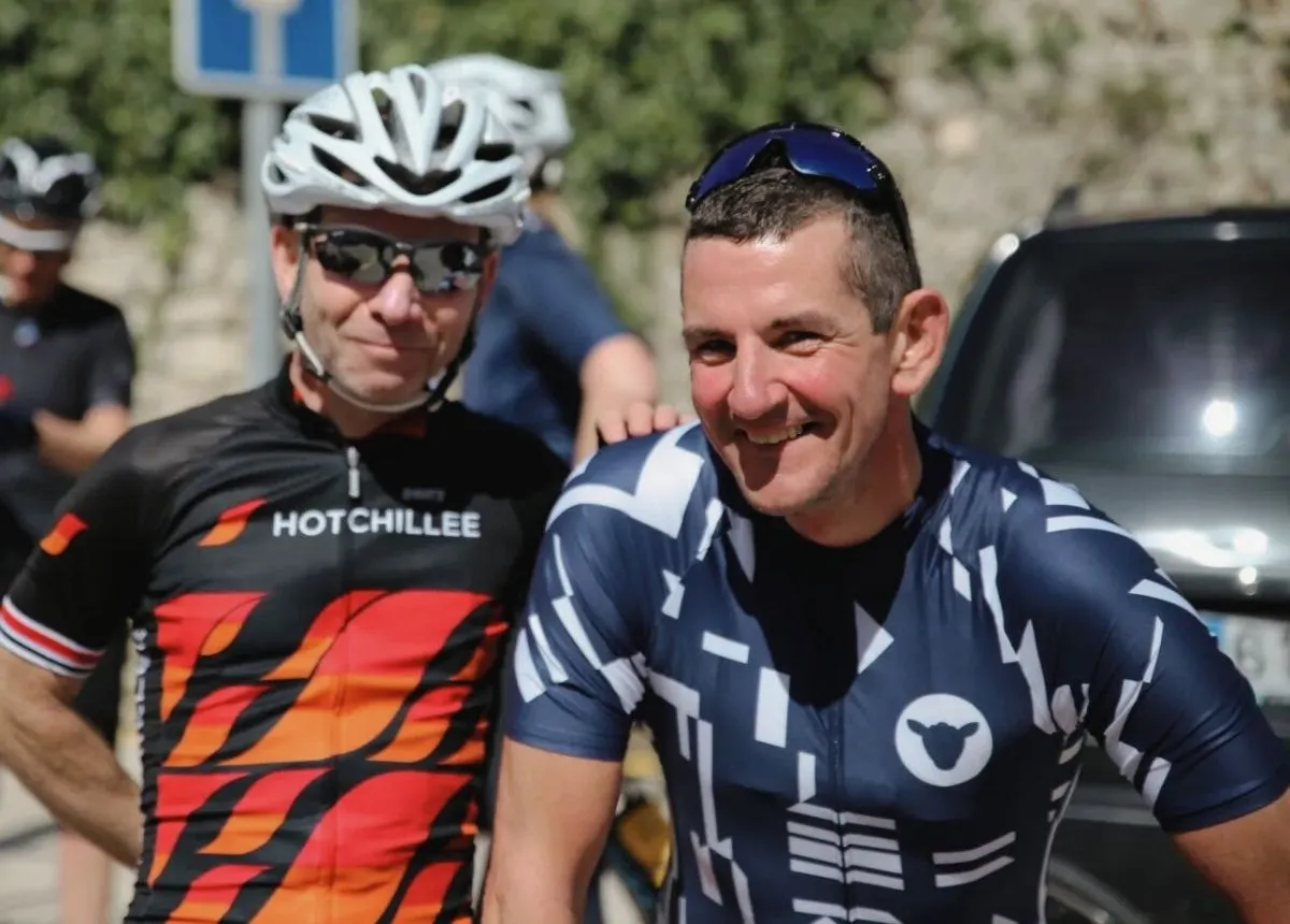 Two cyclists in their forties  in sportswear smiling outdoors, one in black and red HOTCHILLEE jersey and helmet, the other in blue patterned jersey with sunglasses on his head.