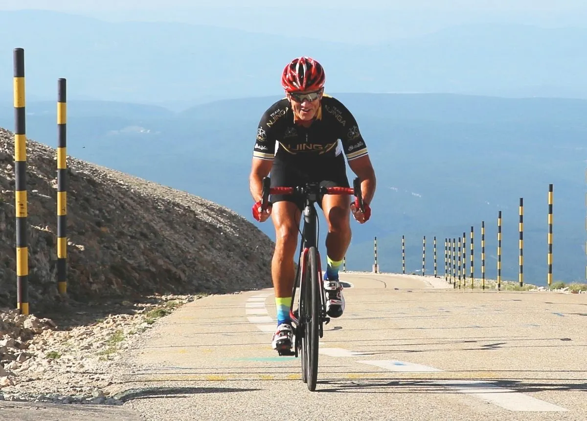 Mature cyclist wearing a red helmet and black cycling outfit riding uphill on a mountain road with black and yellow striped poles.