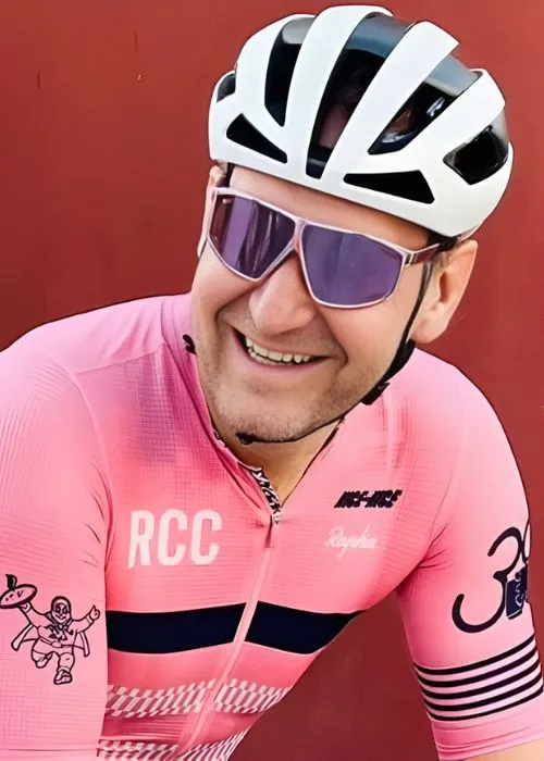 Smiling cyclist over 50 wearing a white and black helmet, reflective sunglasses, and a pink cycling jersey with logos against a red background.