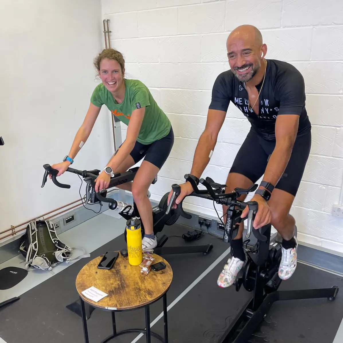Two cyclists on indoor smart bikes in a studio, smiling during a training session.