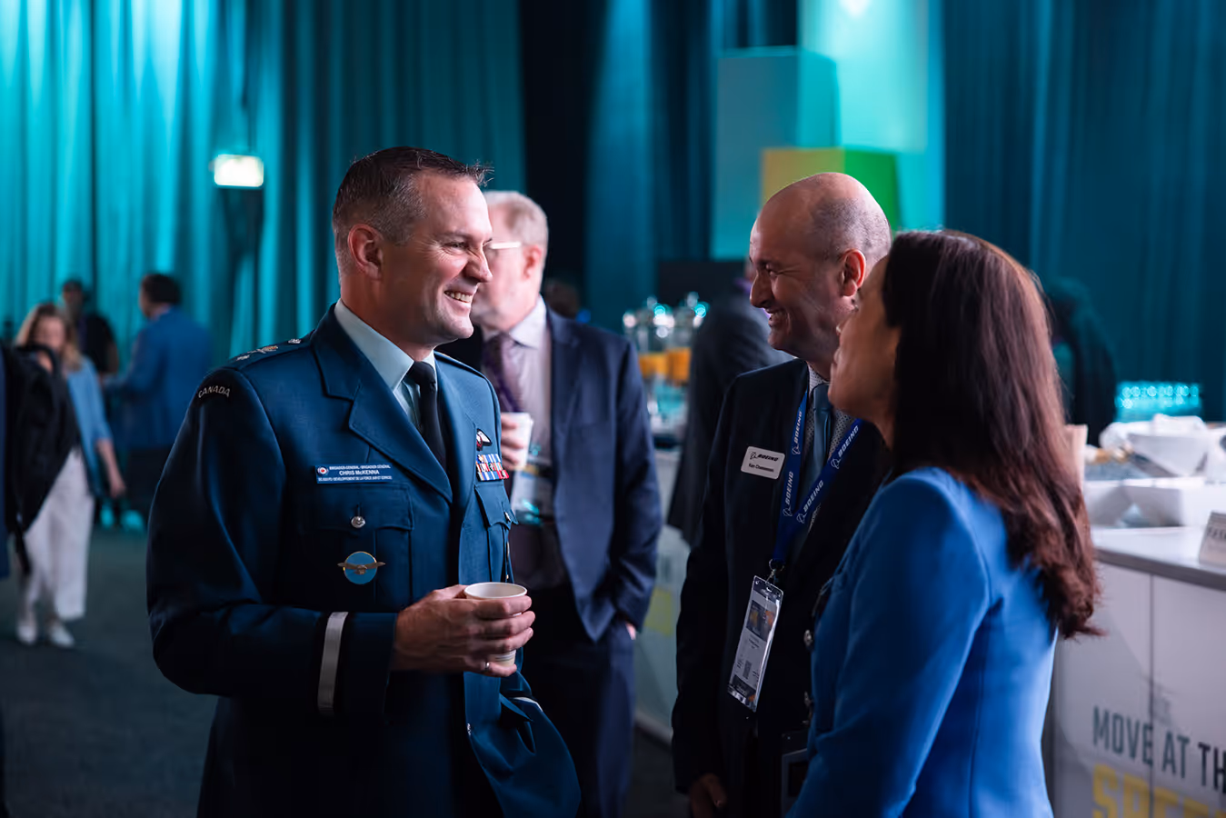 Procurement officers in formal attire speaking at a defence industry event, with exhibition booths visible in the background.