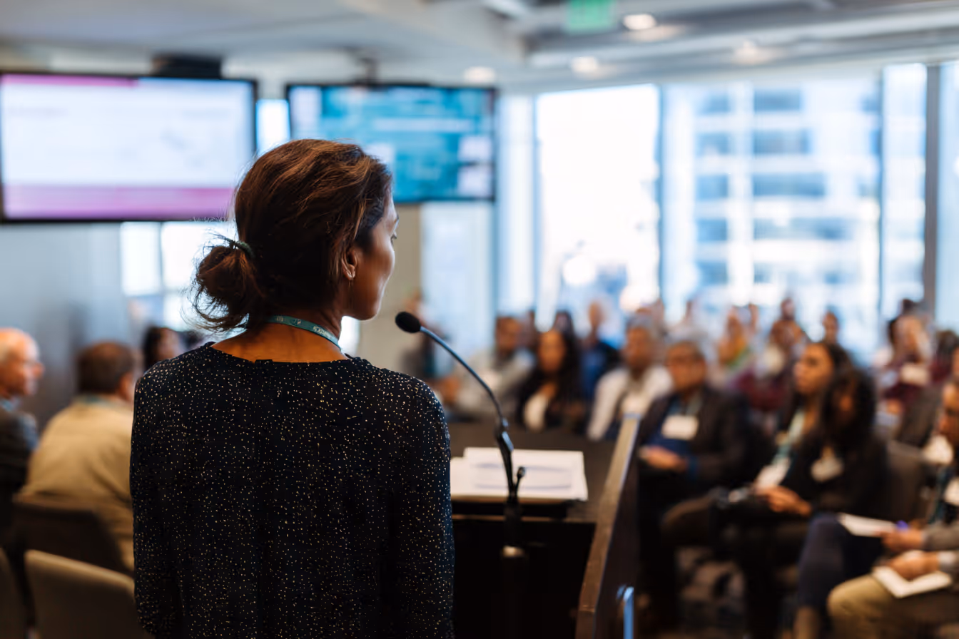 Woman presenting to a large audience in a modern boardroom during an industry intelligence briefing.