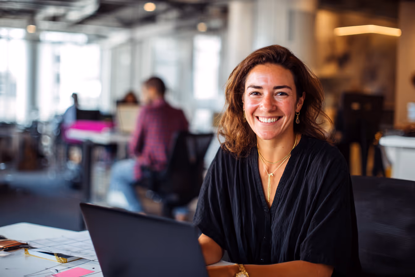 Smiling woman seated at a desk with a laptop in a modern office environment, representing CADSI’s communications team.