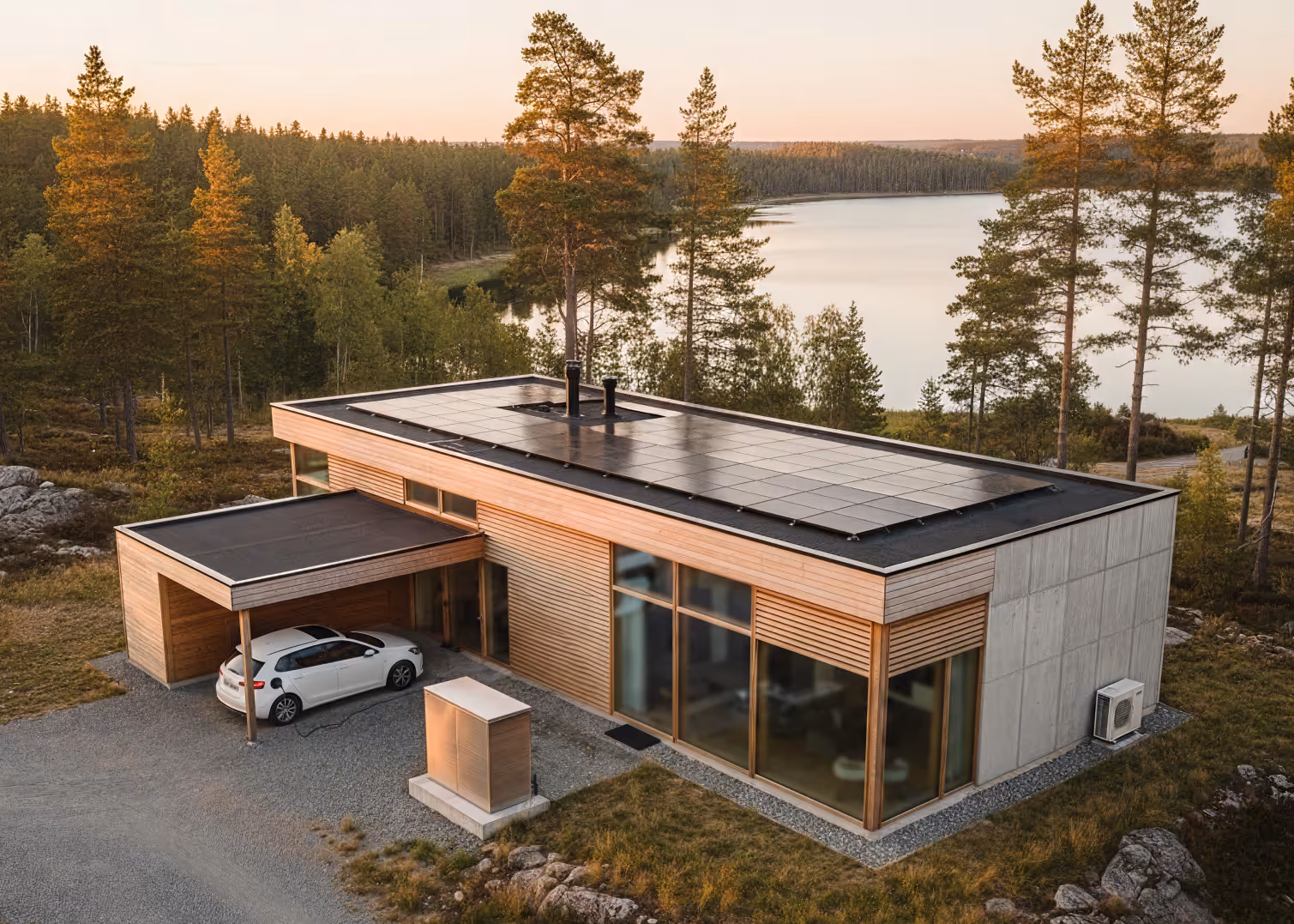 Modernt hus med solpaneler på taket, hemmabatteri och elbil vid sjö i solnedgång