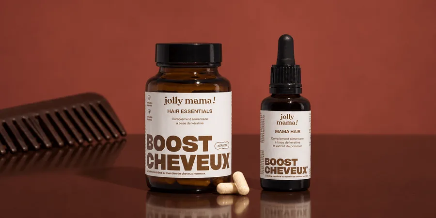 Le pot Hair essentials et le flacon à pipette Mama hair de Jolly Mama, avec deux gélules et un peigne sur un fond brun rouge.