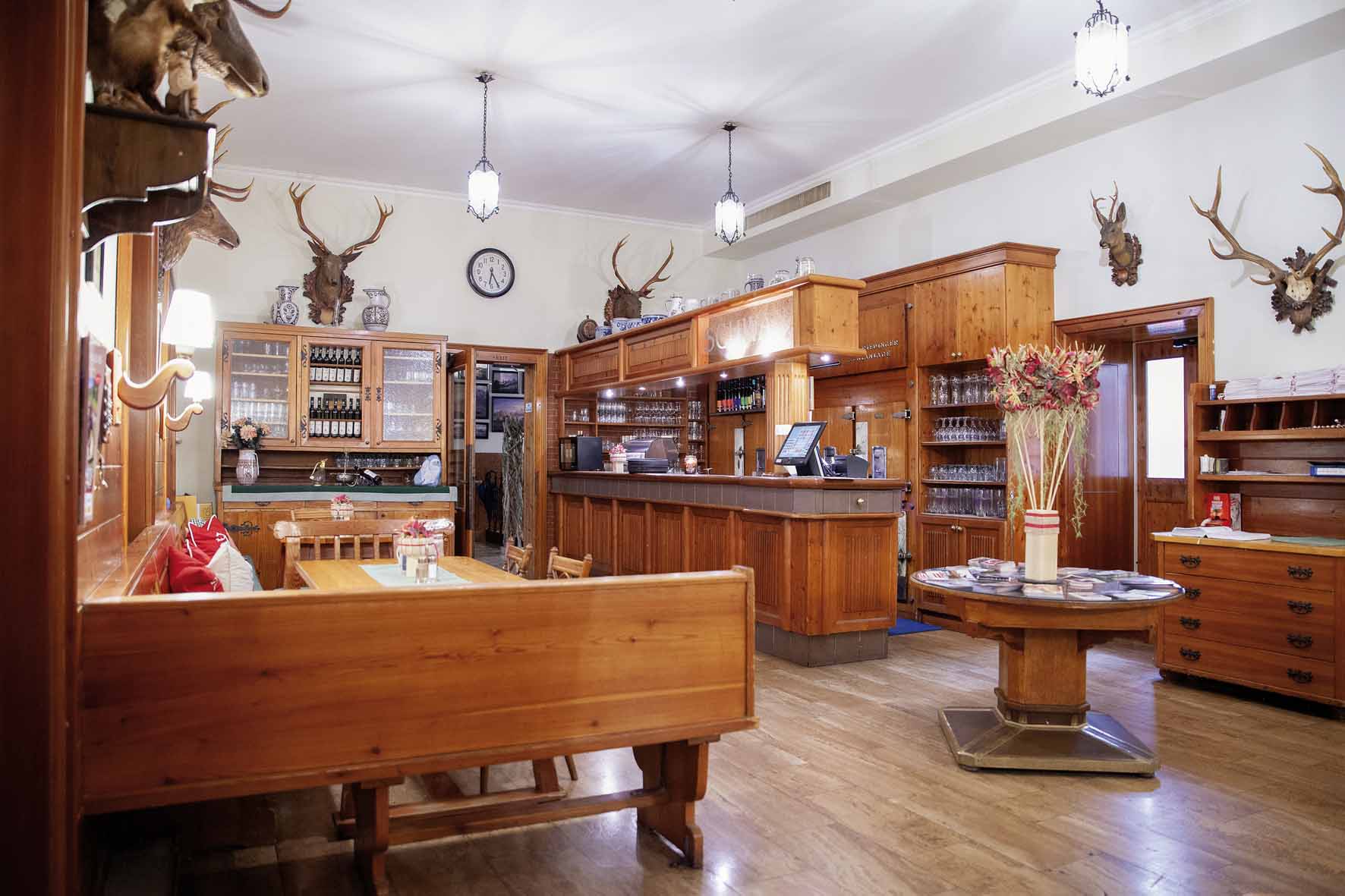 Holzgasthaus mit Tresen, Holztischen, Hirschgeweihen an den Wänden und einer Uhr über einer Vitrine.